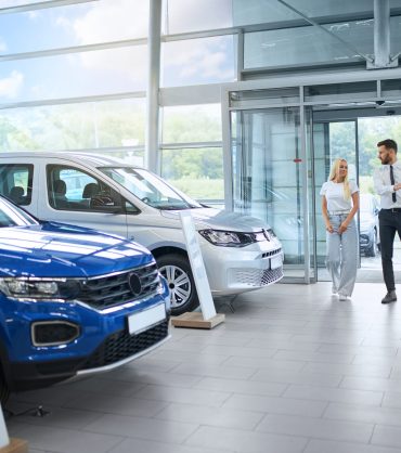 Competent salesman walking with female client at modern auto salon and presenting various luxury car. Young caucasian woman choosing new vehicle at showroom.